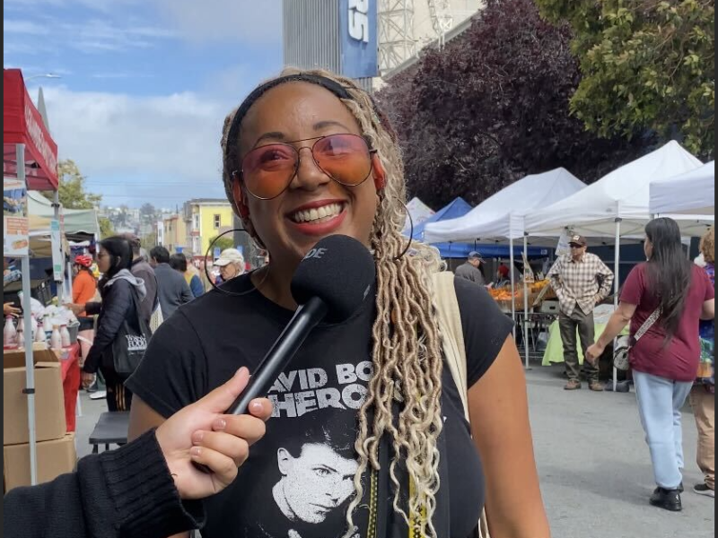 A smiling woman wearing sunglasses and a "David Bowie Heroes" t-shirt is being interviewed at an outdoor market with vendor tents and people in the background.
